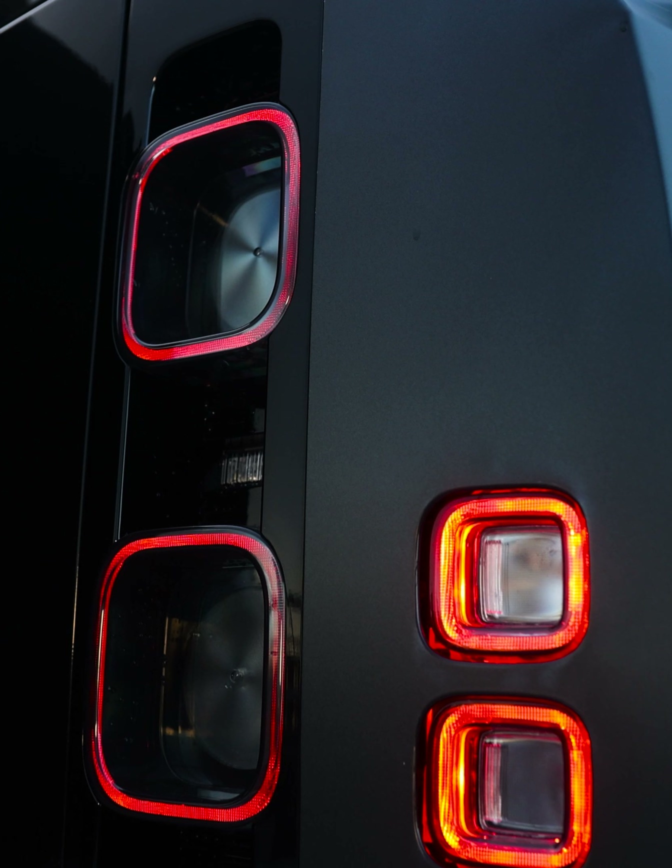 A custom Branders Land Rover Defender 130, in black, is parked with its rear facing a brick building with windows.