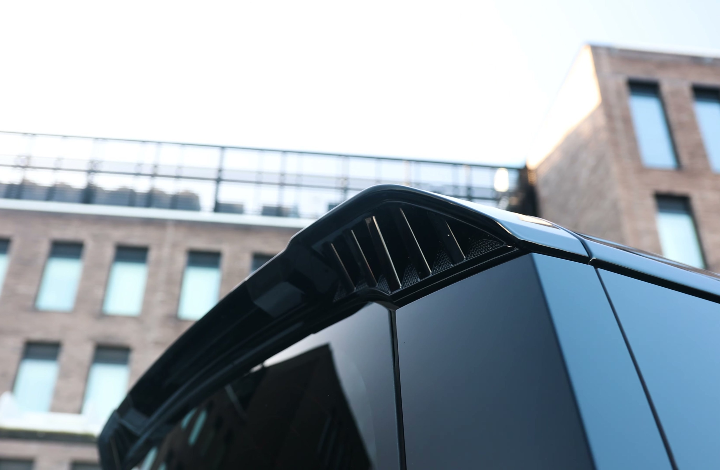 A custom Branders Land Rover Defender 130 with a matte black finish and roof-mounted cargo box is parked in front of a brick building.
