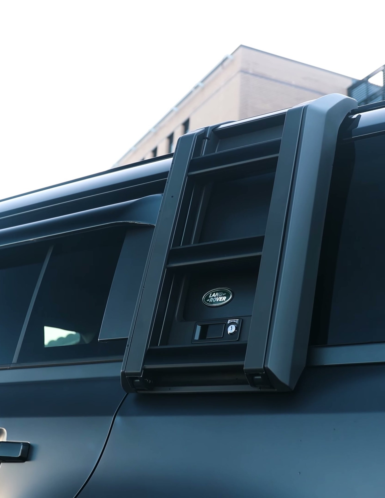 A custom Branders Land Rover Defender 130 with roof-mounted lights and skis, parked in front of a modern brick building.