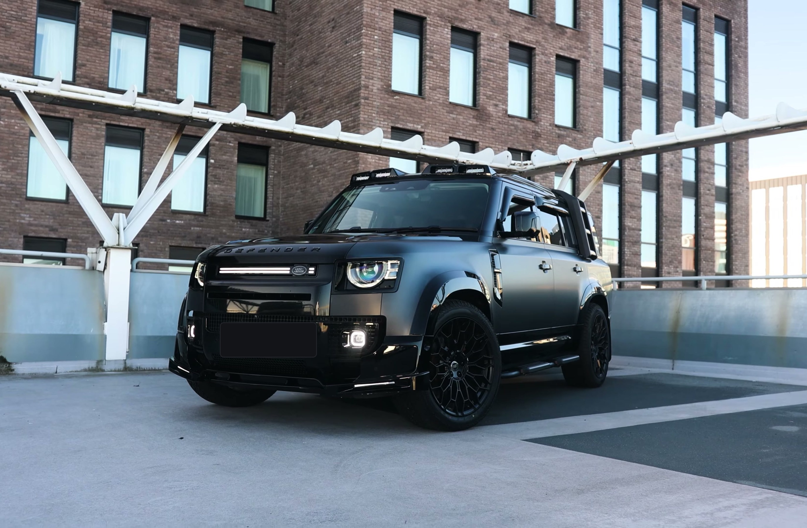 A custom Branders Land Rover Defender 130 in matte black is parked on a concrete rooftop with a brick building in the background.